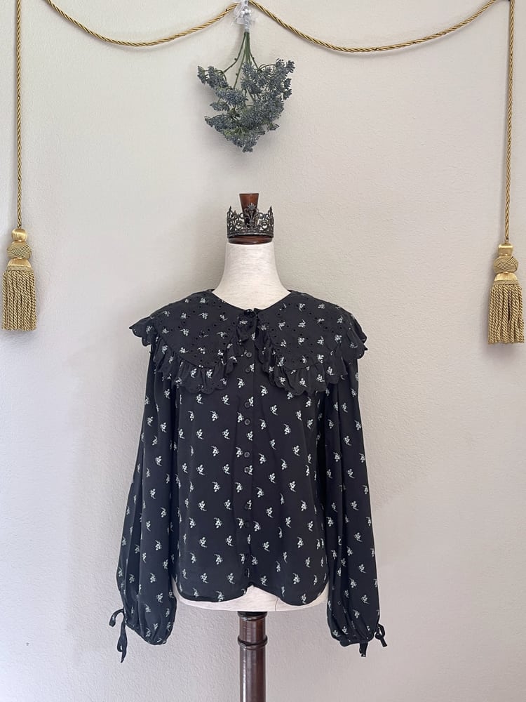 A black floral print historically inspired baroque blouse with large lace trimmed collar and long bishop sleeves is pictured on a mannequin from the front.