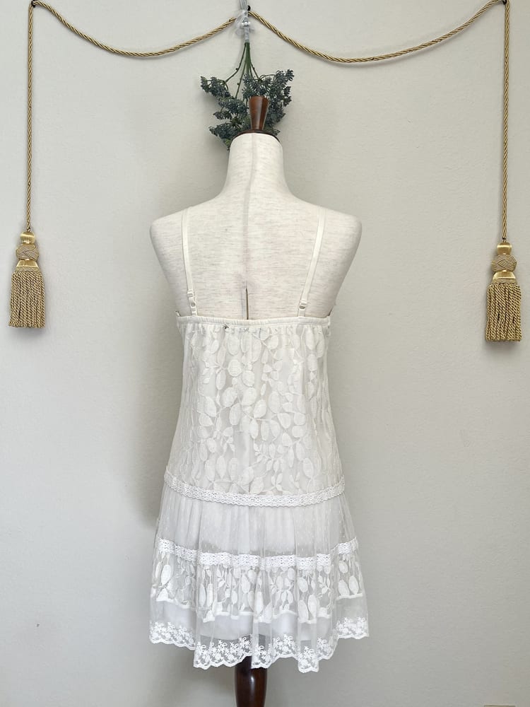 A white y2k crochet and lace fantasy inspired tunic mini dress is pictured on a mannequin from the back.