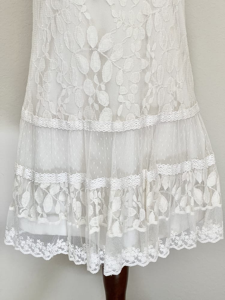 A white y2k crochet and lace fantasy inspired tunic mini dress is pictured on a mannequin from the front.