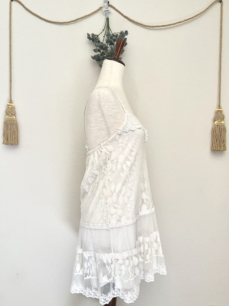 A white y2k crochet and lace fantasy inspired tunic mini dress is pictured on a mannequin from the side.