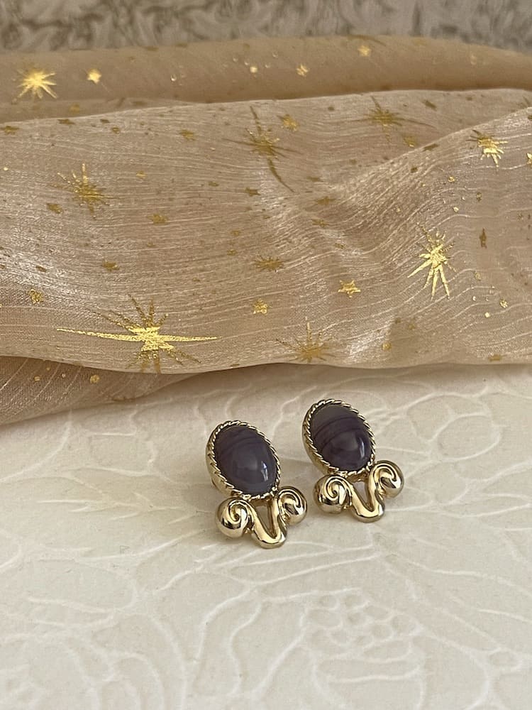 A pair of Historically Inspired Golden Filigree Earrings in Purple Chalcedony stone, on a floral puff with celestial fabric background.