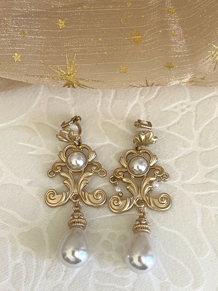 A pair of gold-colored Renaissance Inspired Ornate Flourish Earrings with Pearl Drops, pictured on a floral puff.