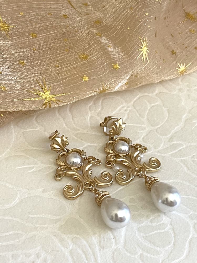 A pair of gold-colored Renaissance Inspired Ornate Flourish Earrings with Pearl Drops, pictured on a floral puff.