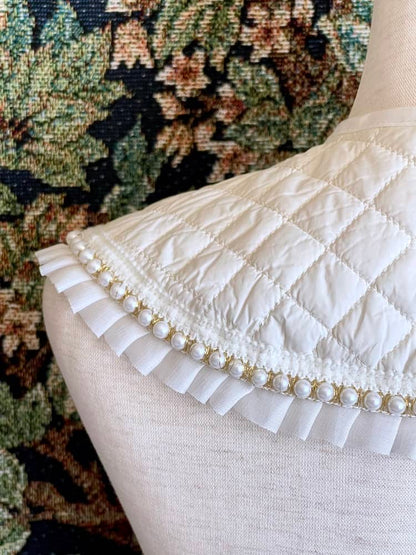 A Historically Inspired Quilted Round Collar with Ruffle & Pearl Trim in Ivory/White pictured on a mannequin in front of a historical tapestry.