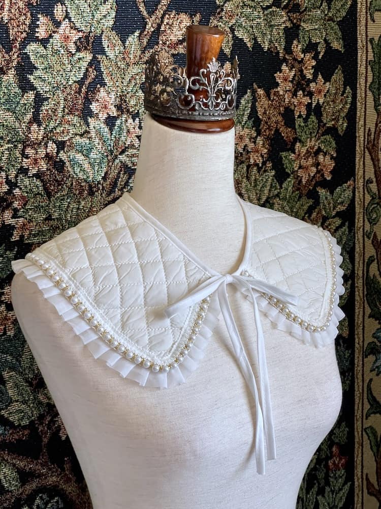 A Historically Inspired Quilted Round Collar with Ruffle & Pearl Trim in Ivory/White pictured on a mannequin in front of a historical tapestry.
