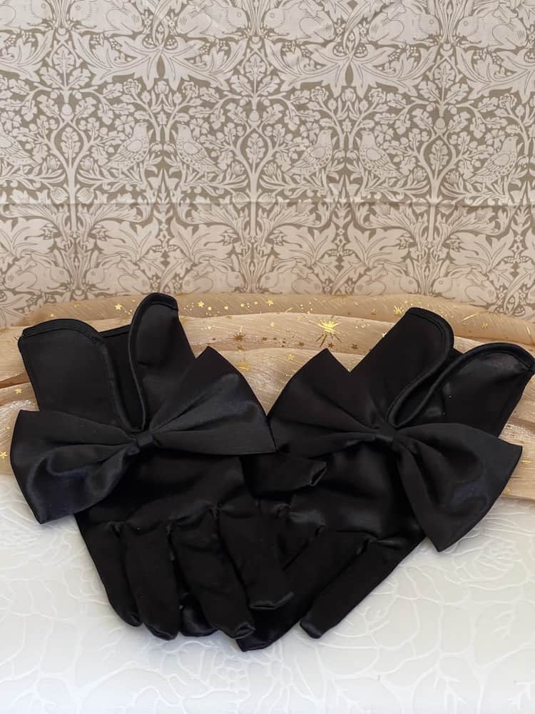 A pair of historically inspired Regency & Victorian era Satin Gloves with Large Bow Detail in Black, pictured on a historical backdrop.