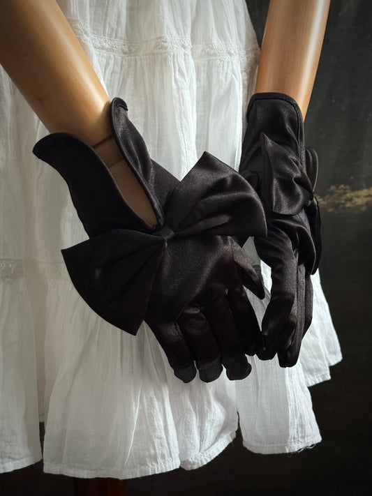 A pair of historically inspired Regency & Victorian era Satin Gloves with Large Bow Detail in Black, pictured on a mannequin in front of an art historical backdrop.