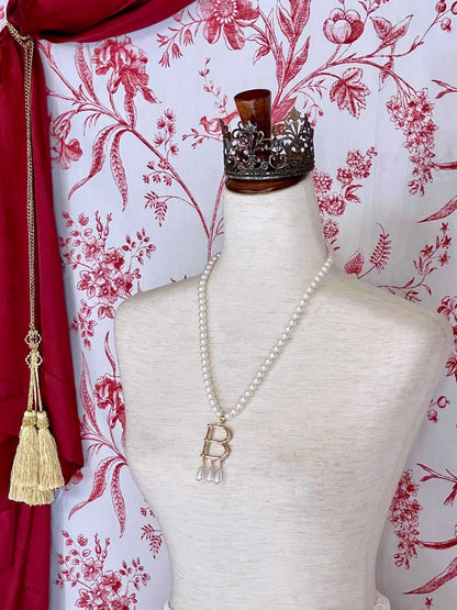 A Historical Tudor Fashion Inspired Anne Boleyn "B" Pearl Necklace on a mannequin in front of an ornate backdrop. 