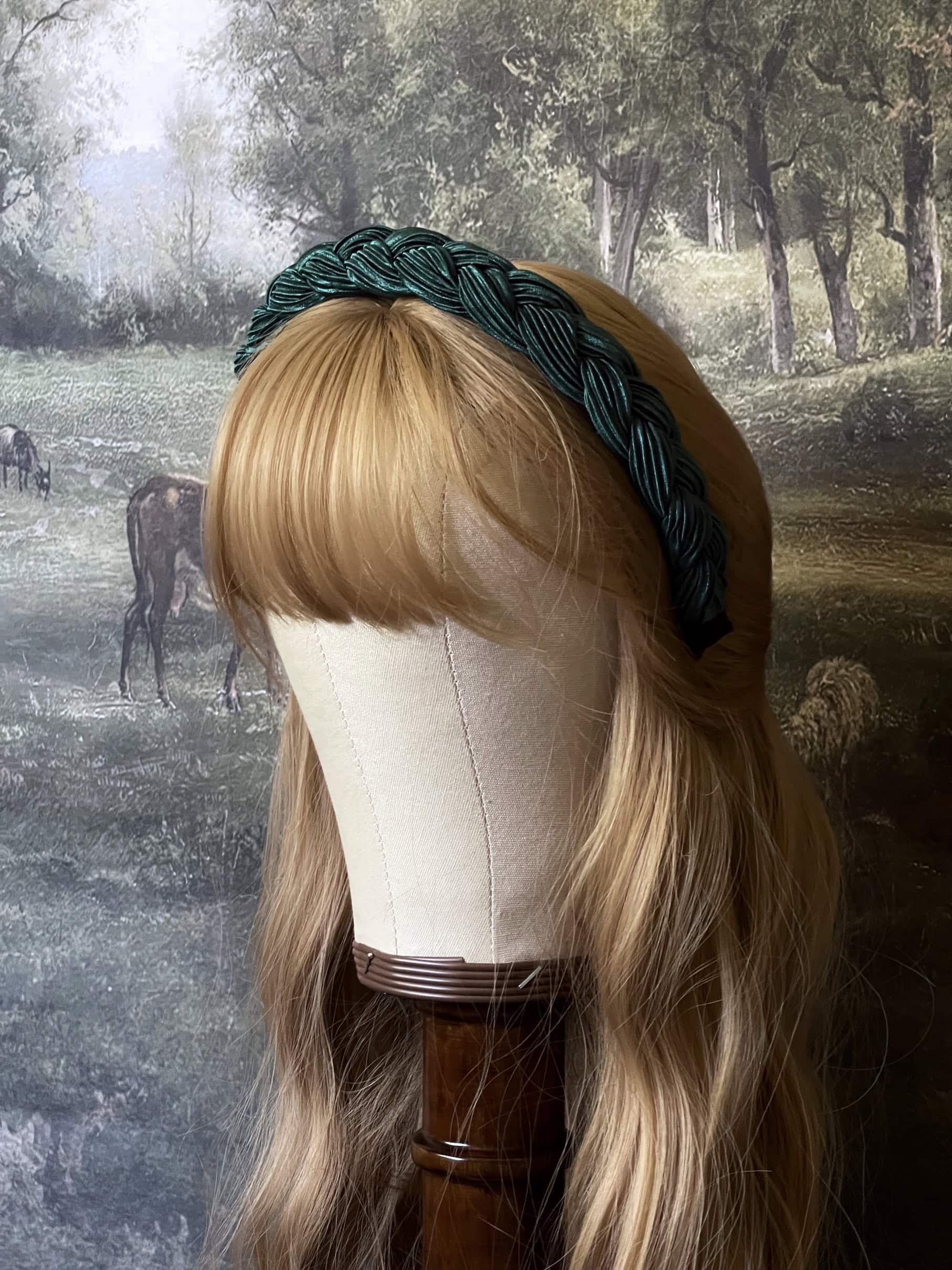 A historically inspired vintage style braided headband in dark forest green is pictured on a mannequin in front of an art history backdrop.