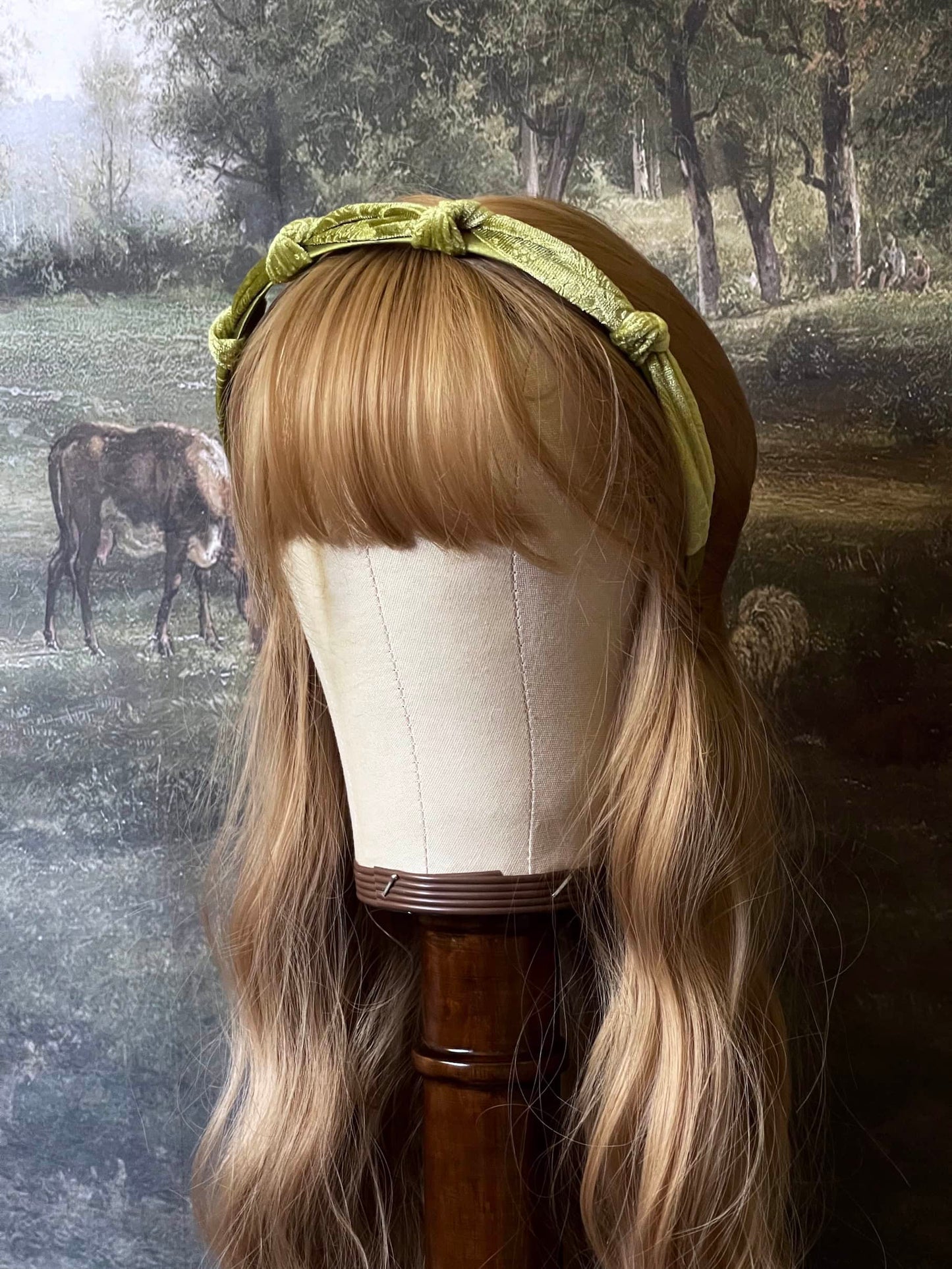 A historically inspired vintage style velvet floral knotted headband in mossy olive green is pictured on a mannequin in front of an art history backdrop.