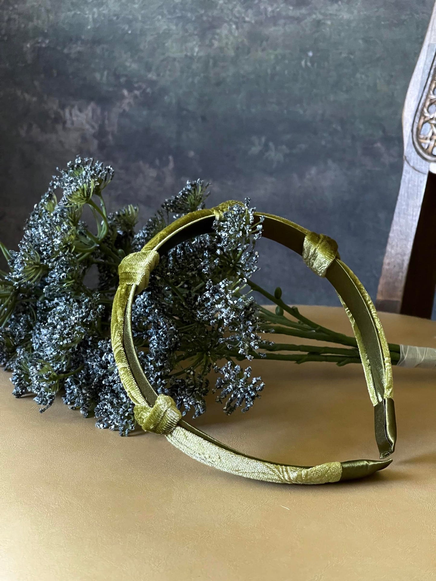 A historically inspired vintage style velvet floral knotted headband in mossy olive green is pictured with a bouquet in front of an art history backdrop.