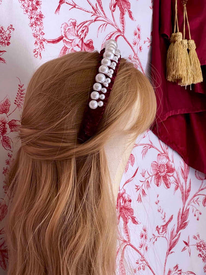 A Historically Inspired French Hood Style Velvet Pearl Headband in Burgundy in front of a toile backdrop.