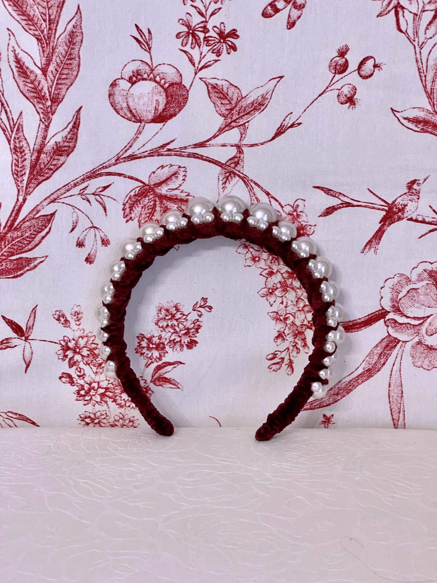 A Historically Inspired French Hood Style Velvet Pearl Headband in Burgundy in front of a toile backdrop.