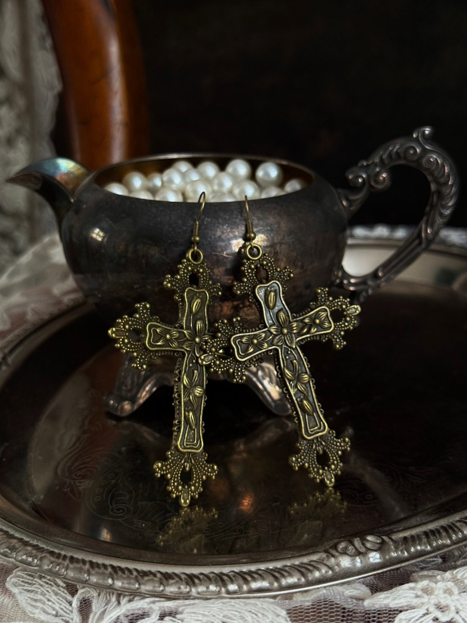 A pair of antique gold / bronze metal cross earrings with ornate floral detailing, hanging on an antique silver cup.
