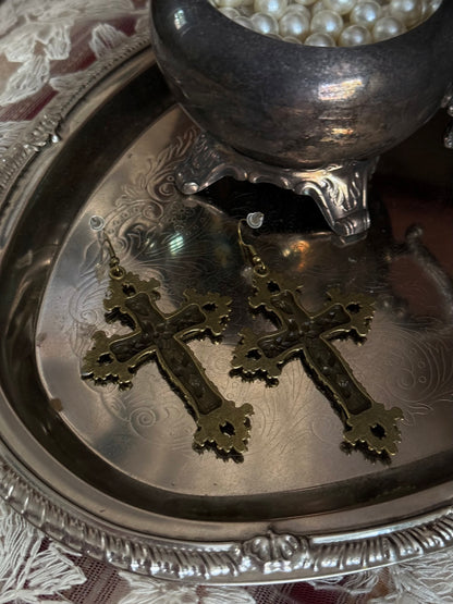 A pair of antique gold / bronze metal cross earrings with ornate floral detailing, sitting on a silver tray