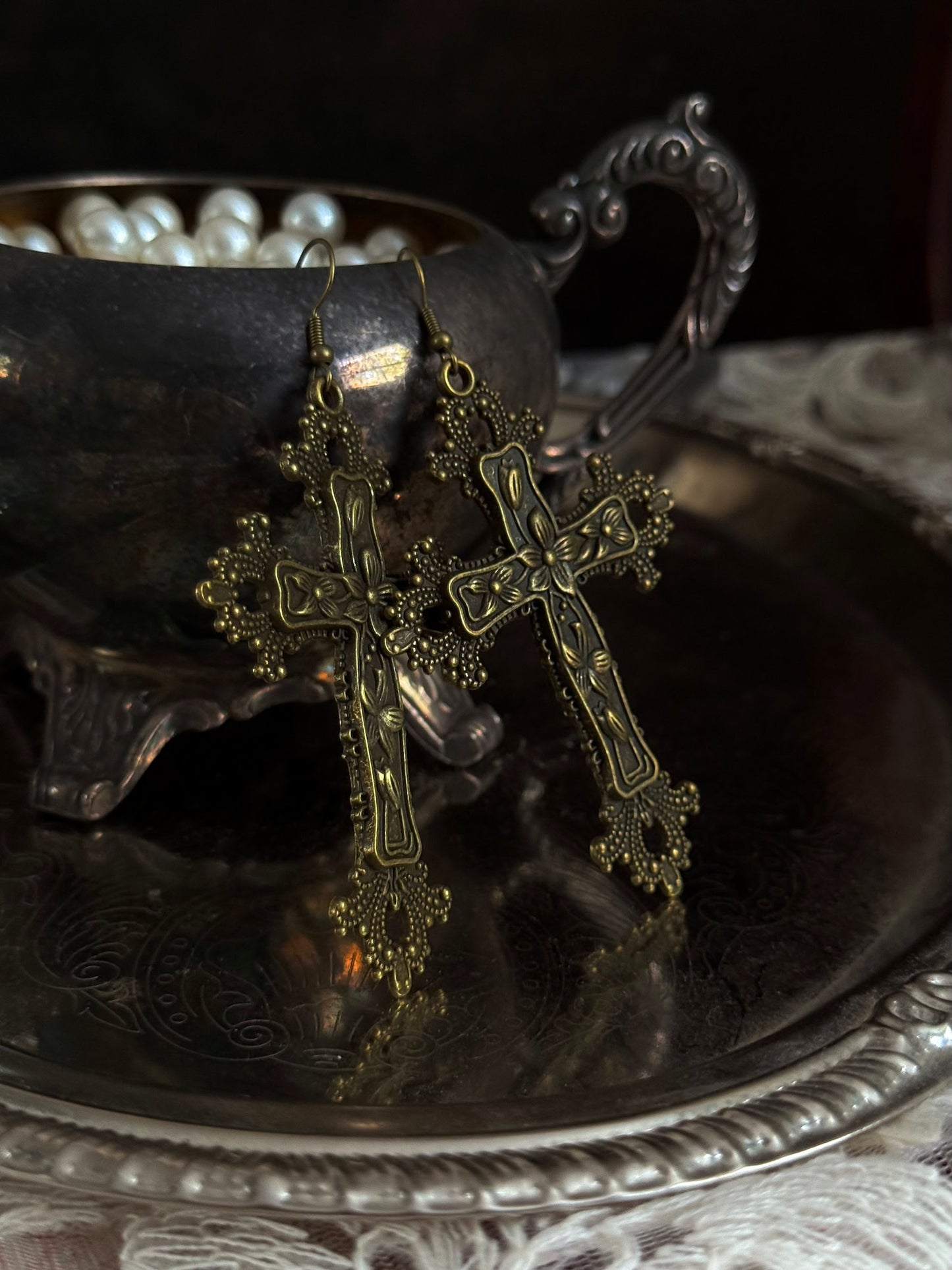 A pair of antique gold / bronze metal cross earrings with ornate floral detailing, hanging on an antique silver cup.