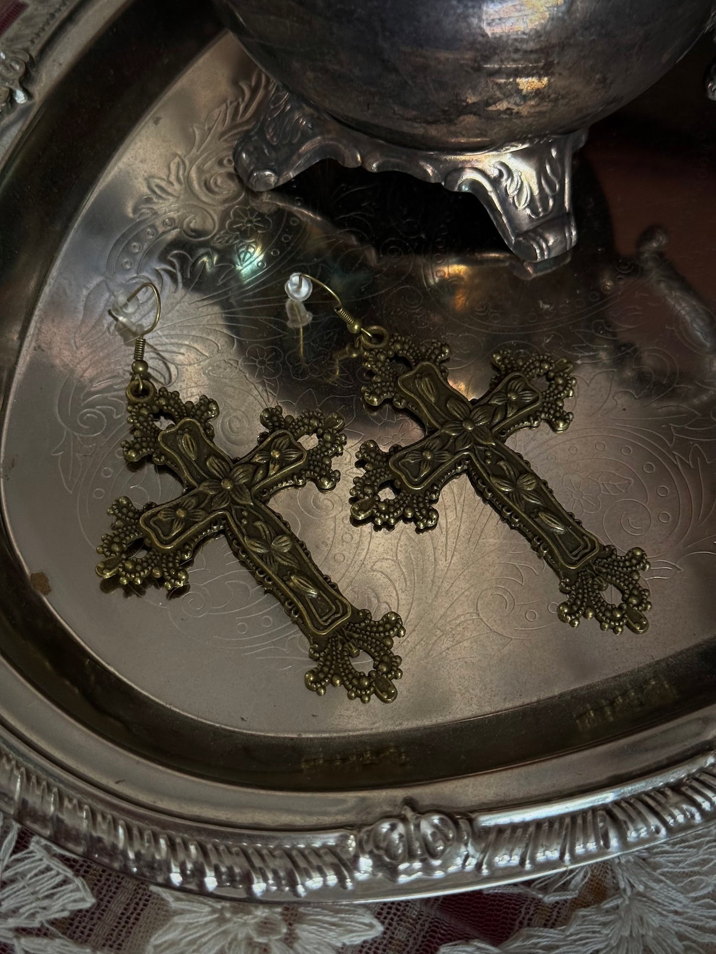 A pair of antique gold / bronze metal cross earrings with ornate floral detailing, sitting on a silver tray