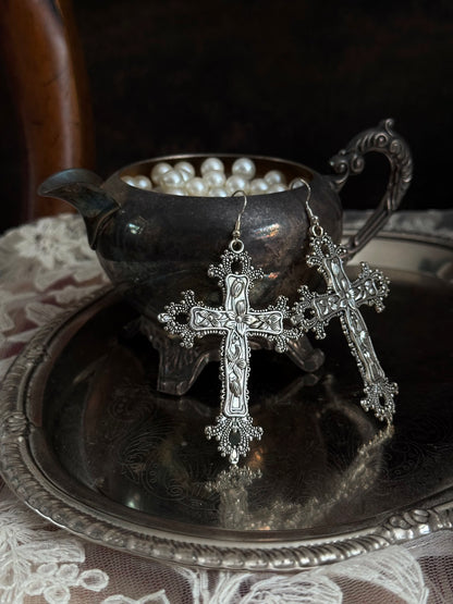 A pair of silver colored metal cross earrings with ornate floral detailing, hanging from an antique silver cup