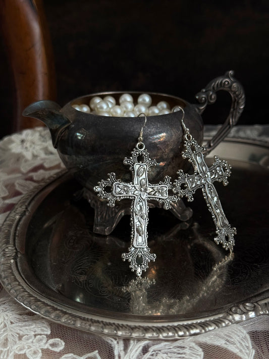 A pair of silver colored metal cross earrings with ornate floral detailing, hanging from an antique silver cup