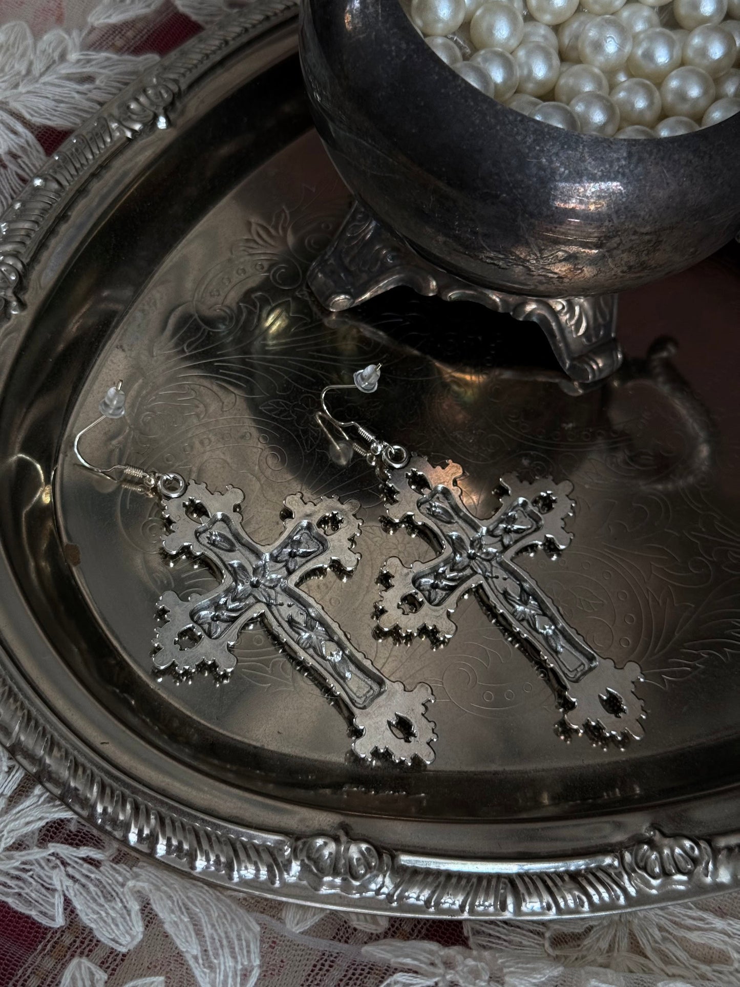 A pair of silver colored metal cross earrings with ornate floral detailing, sitting on a silver tray