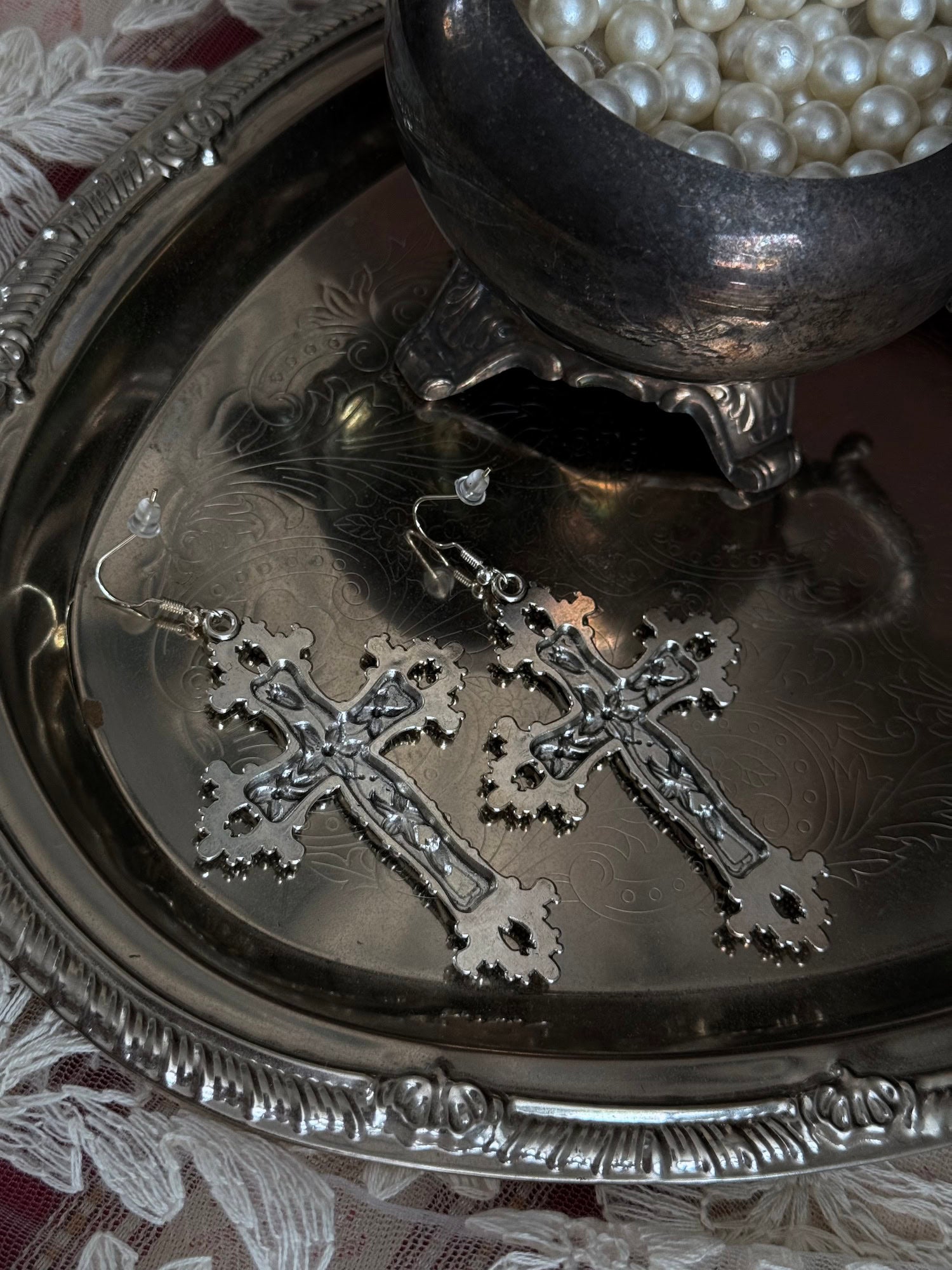 A pair of silver colored metal cross earrings with ornate floral detailing, sitting on a silver tray