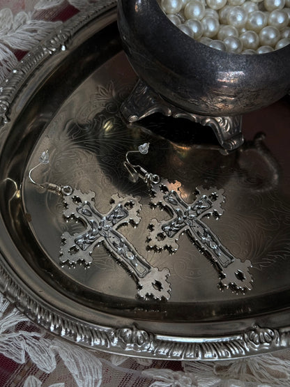 A pair of silver colored metal cross earrings with ornate floral detailing, sitting on a silver tray