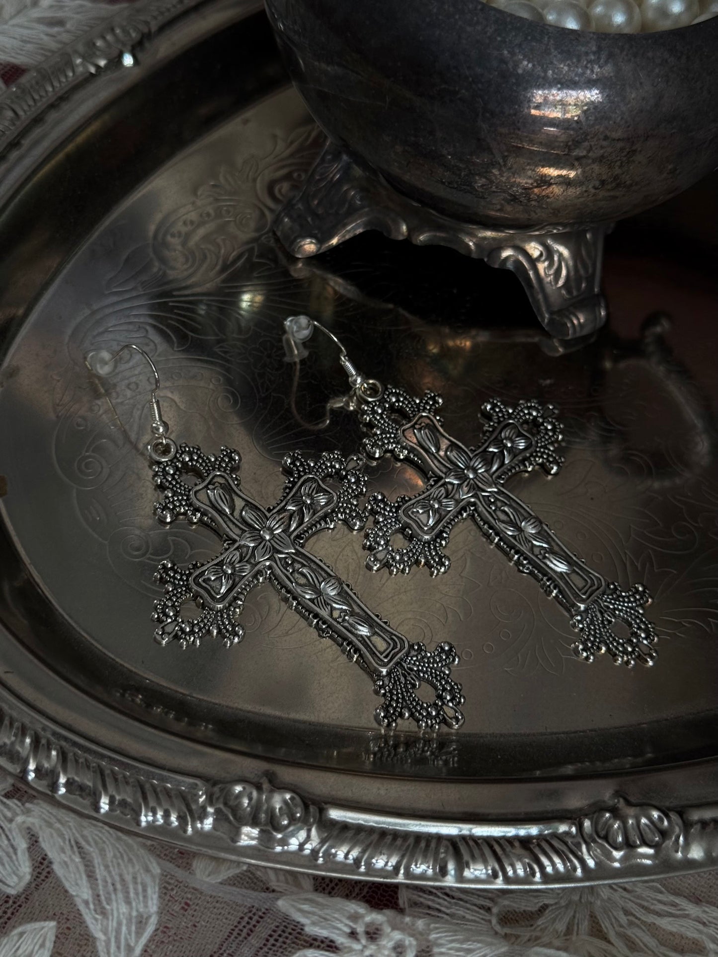 A pair of silver colored metal cross earrings with ornate floral detailing, sitting on a silver tray