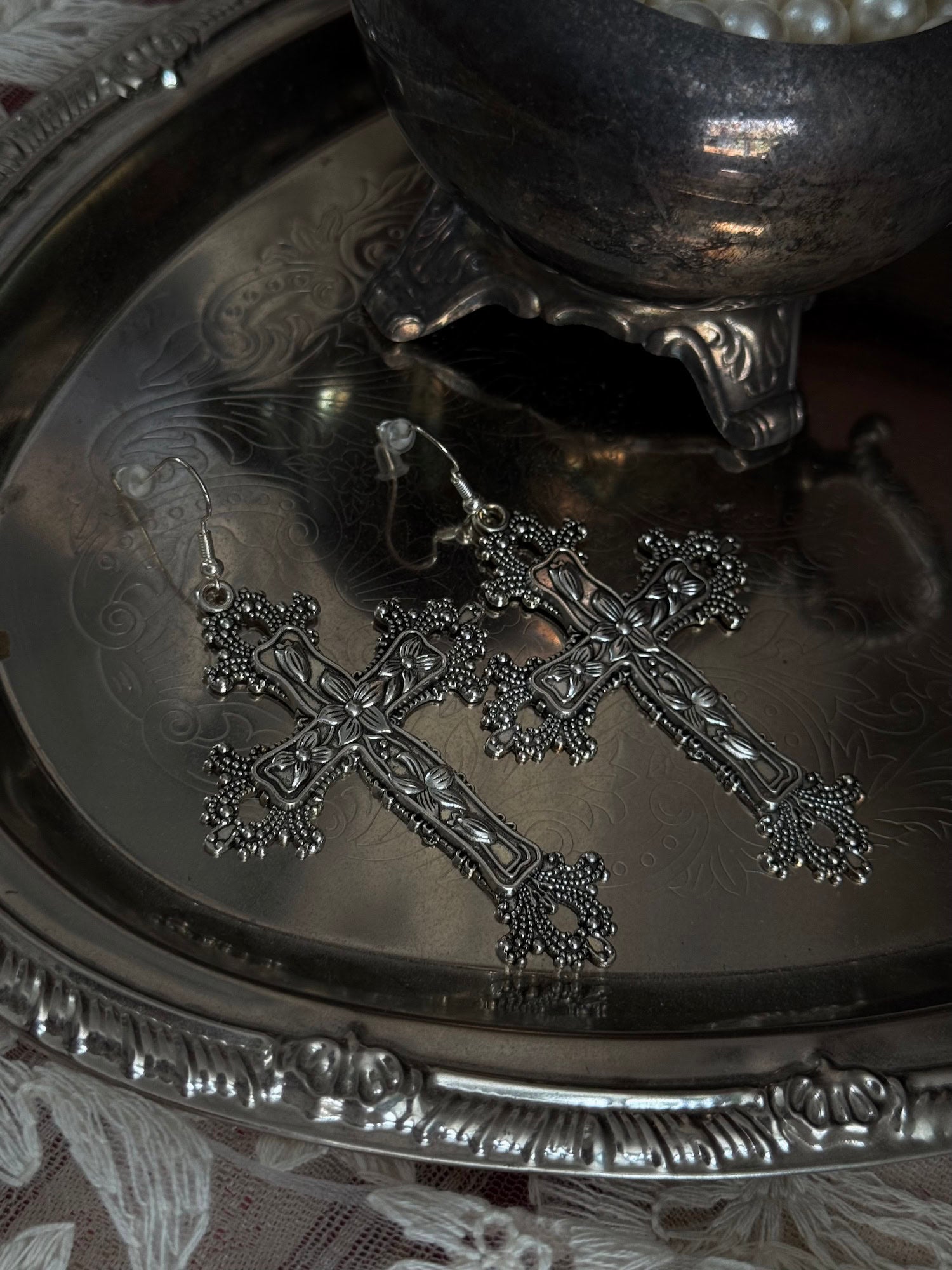 A pair of silver colored metal cross earrings with ornate floral detailing, sitting on a silver tray