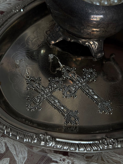 A pair of silver colored metal cross earrings with ornate floral detailing, sitting on a silver tray