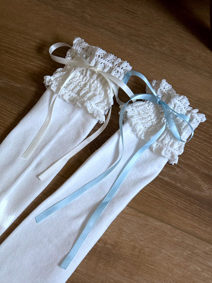 Two pairs of Historically-inspired vintage style white knit socks with lace garter style trim and either blue or white satin ribbons on a wooden floor.