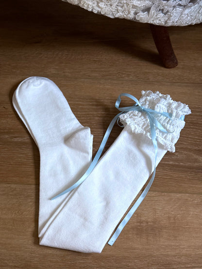 A pair of Historically-inspired vintage style white knit socks with lace garter style trim and blue satin ribbons on a wooden floor.