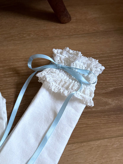 A pair of Historically-inspired vintage style white knit socks with lace garter style trim and blue satin ribbons on a wooden floor.