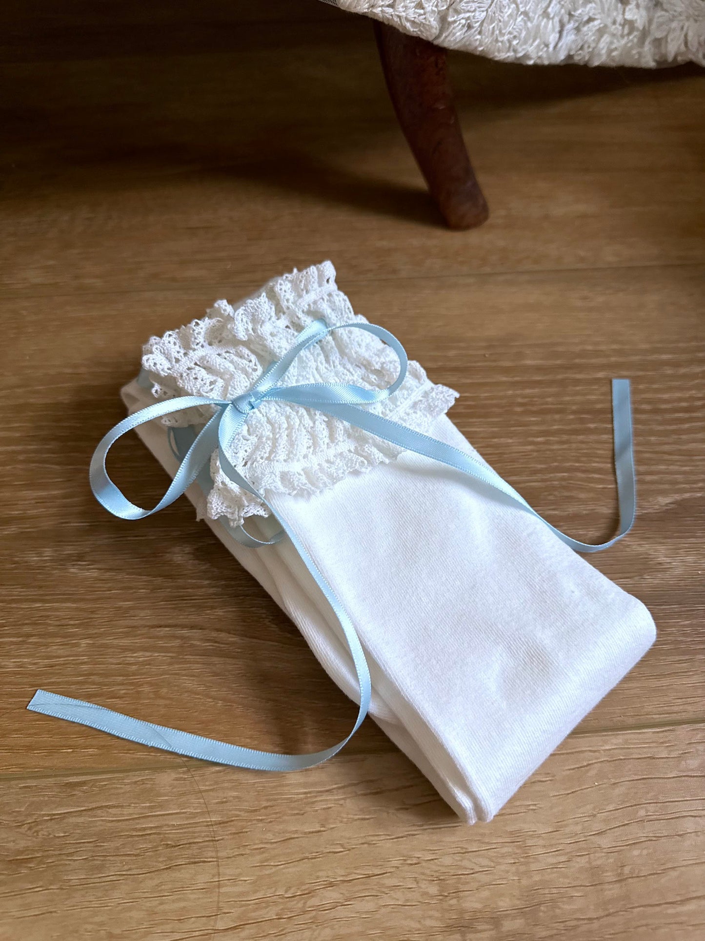 A pair of Historically-inspired vintage style white knit socks with lace garter style trim and blue satin ribbons on a wooden floor.