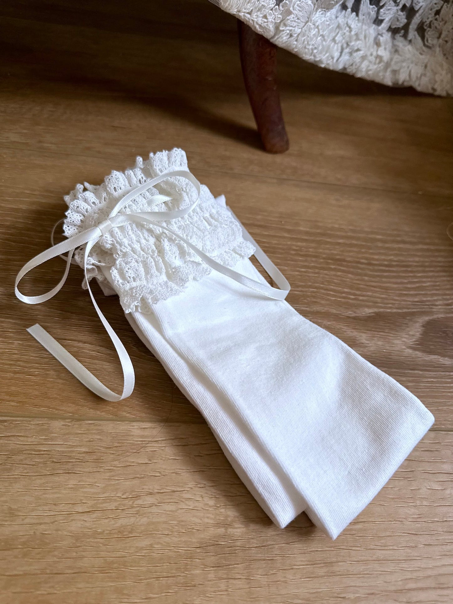 Historically-inspired vintage style white knit socks with lace garter style trim and white satin ribbons on a wooden floor.