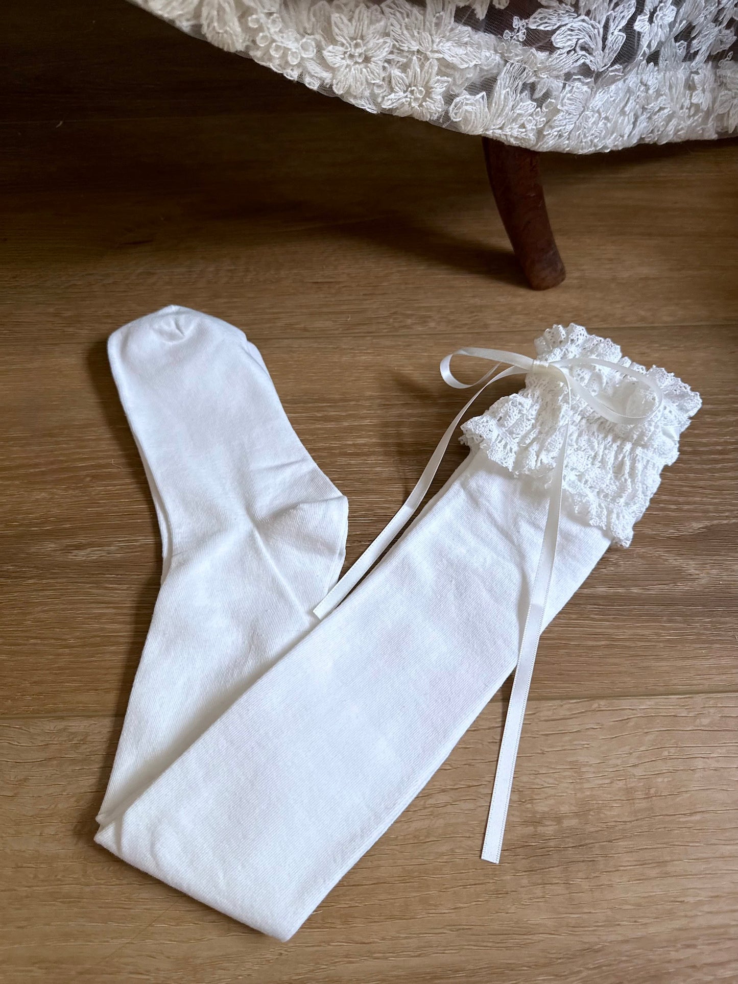 Historically-inspired vintage style white knit socks with lace garter style trim and white satin ribbons on a wooden floor.