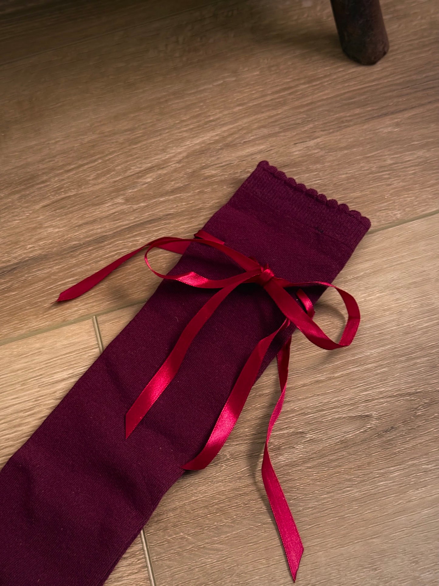 A pair of Historically Inspired Cotton Knee-High Socks with Satin Ribbon Lacing in a dark Burgundy red, pictured on a wooden cottage floor.