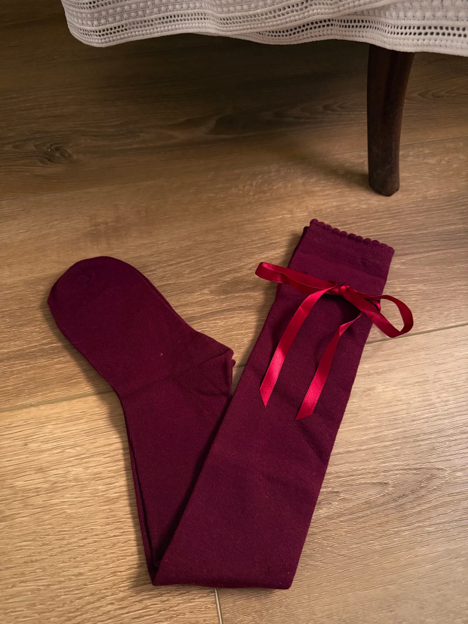A pair of Historically Inspired Cotton Knee-High Socks with Satin Ribbon Lacing in a dark Burgundy red, pictured on a wooden cottage floor.