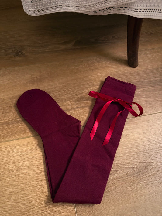 A pair of Historically Inspired Cotton Knee-High Socks with Satin Ribbon Lacing in a dark Burgundy red, pictured on a wooden cottage floor.