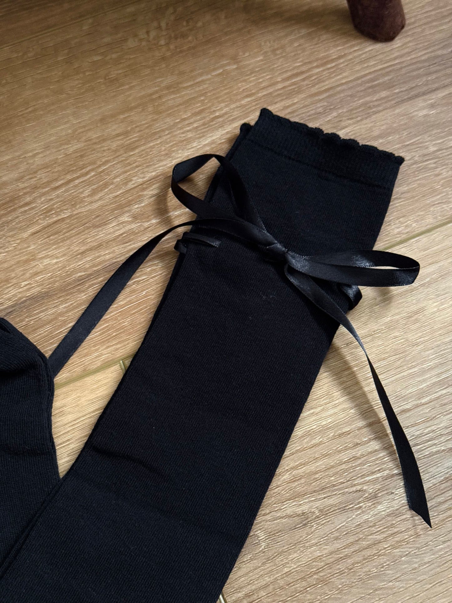 A pair of Historically Inspired Cotton Knee-High Socks with Satin Ribbon Lacing in  Gothic Black, pictured on a wooden cottage floor.