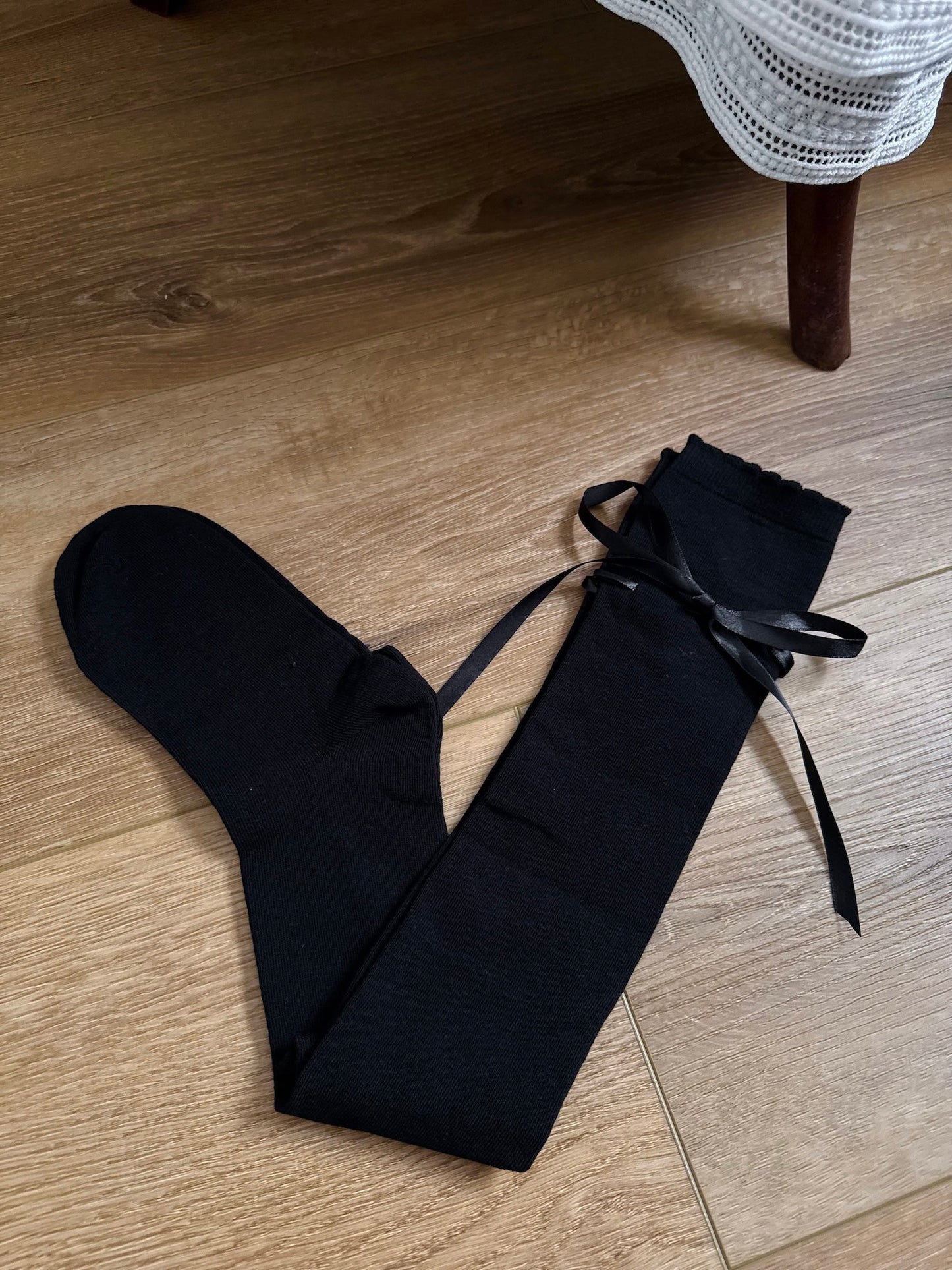 A pair of Historically Inspired Cotton Knee-High Socks with Satin Ribbon Lacing in  Gothic Black, pictured on a wooden cottage floor.