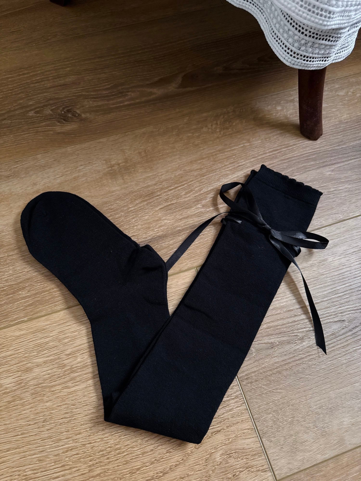 A pair of Historically Inspired Cotton Knee-High Socks with Satin Ribbon Lacing in  Gothic Black, pictured on a wooden cottage floor.