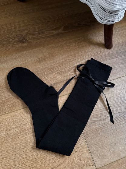 A pair of Historically Inspired Cotton Knee-High Socks with Satin Ribbon Lacing in  Gothic Black, pictured on a wooden cottage floor.