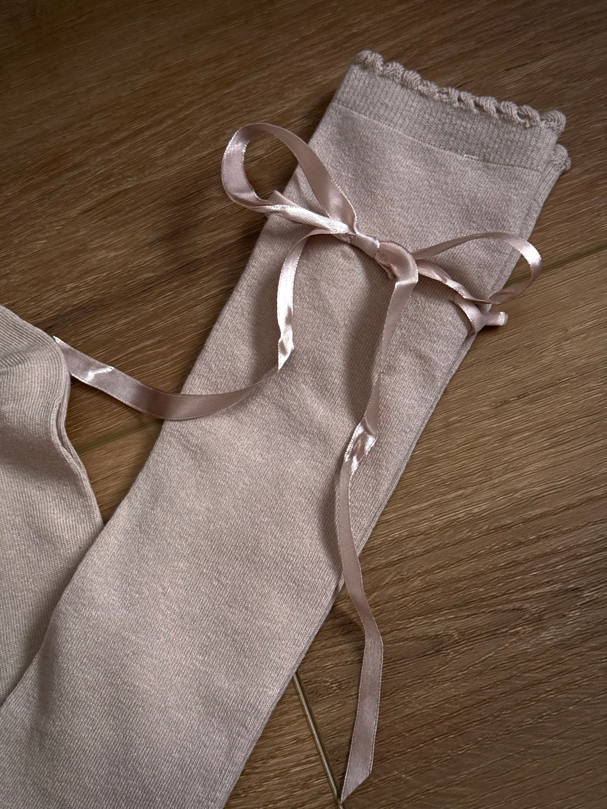 A pair of Historically Inspired Cotton Knee-High Socks with Satin Ribbon Lacing in oatmeal beige, pictured on a historic wooden cottage floor.