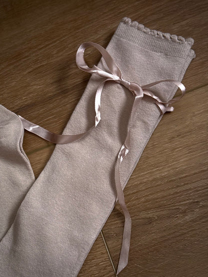 A pair of Historically Inspired Cotton Knee-High Socks with Satin Ribbon Lacing in oatmeal beige, pictured on a historic wooden cottage floor.