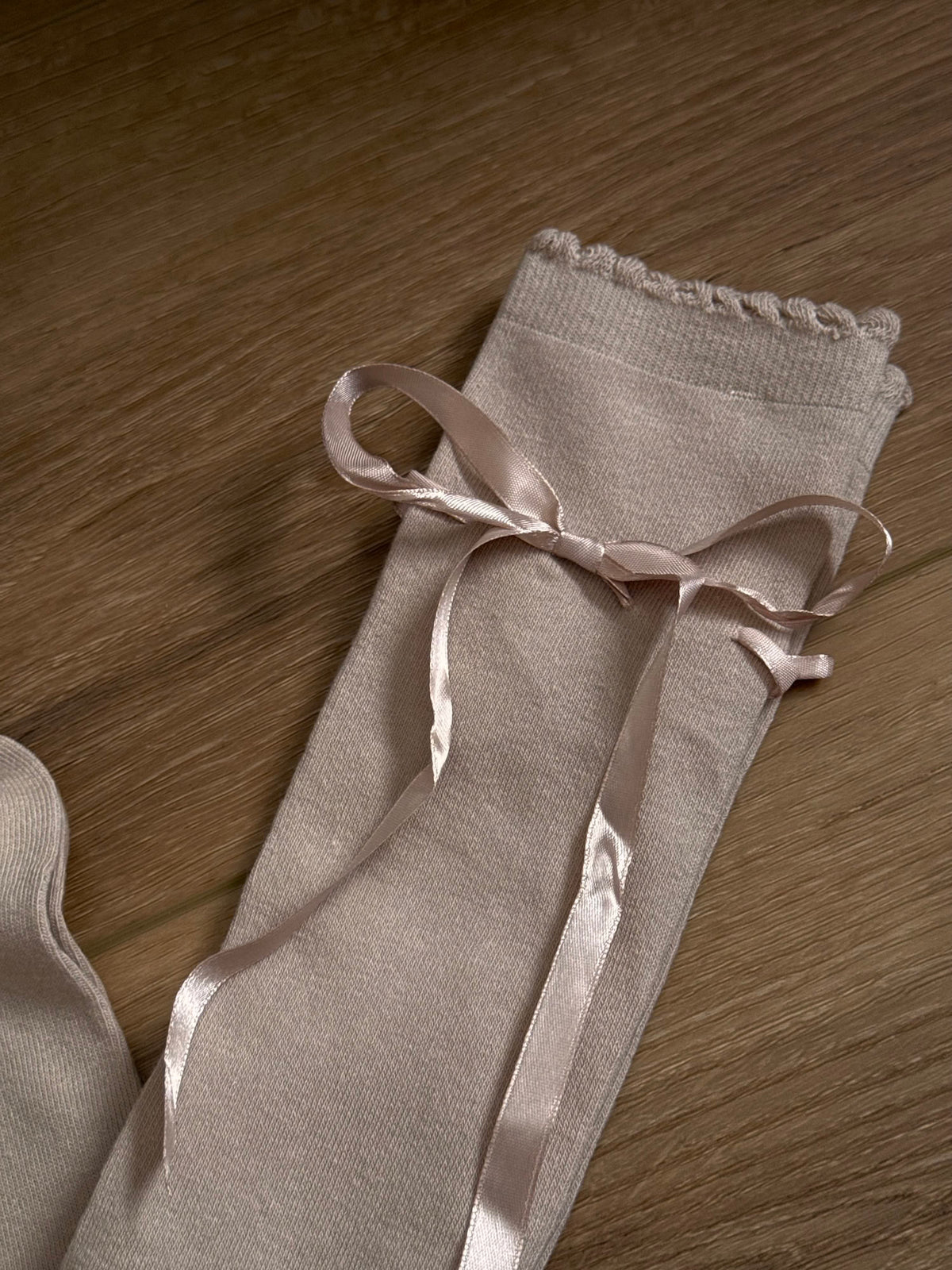 A pair of Historically Inspired Cotton Knee-High Socks with Satin Ribbon Lacing in oatmeal beige, pictured on a historic wooden cottage floor.