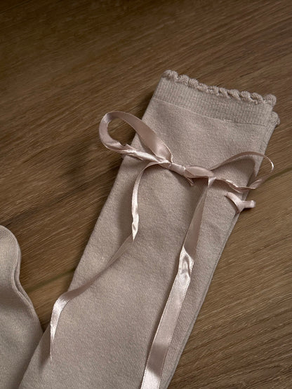 A pair of Historically Inspired Cotton Knee-High Socks with Satin Ribbon Lacing in oatmeal beige, pictured on a historic wooden cottage floor.