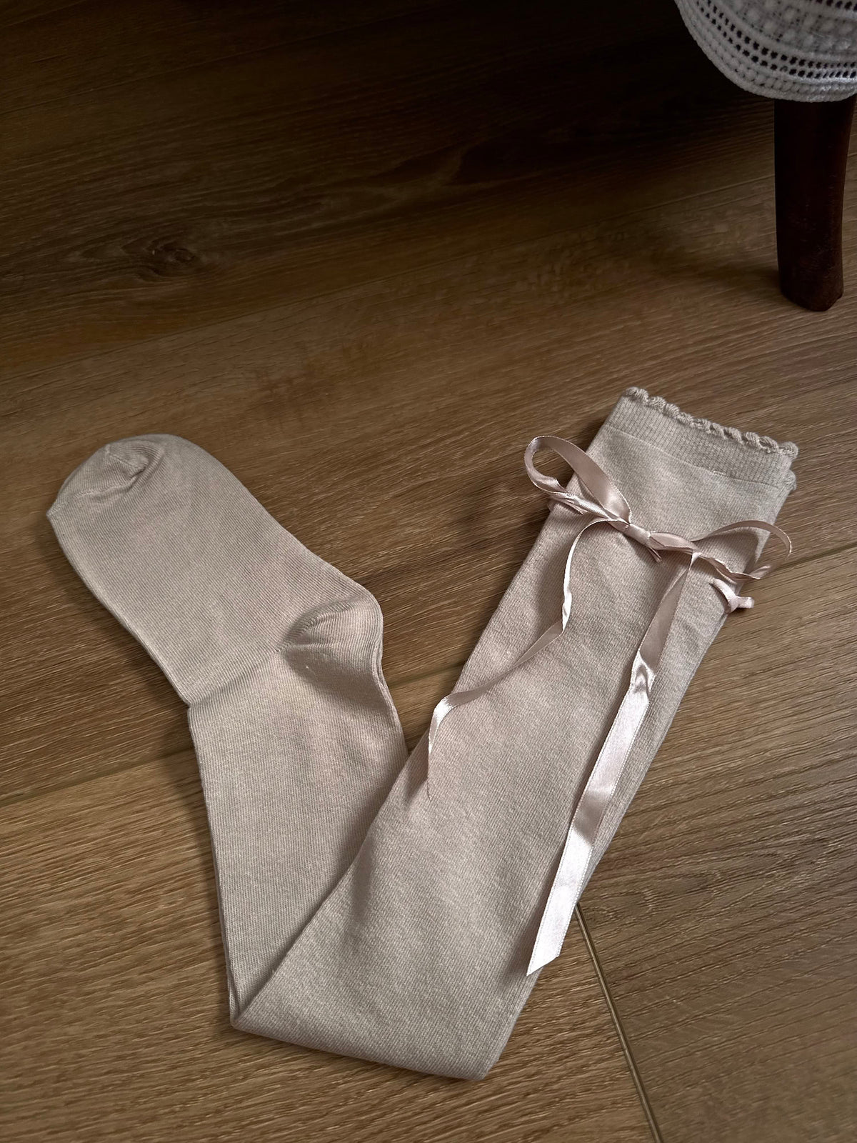 A pair of Historically Inspired Cotton Knee-High Socks with Satin Ribbon Lacing in oatmeal beige, pictured on a historic wooden cottage floor.