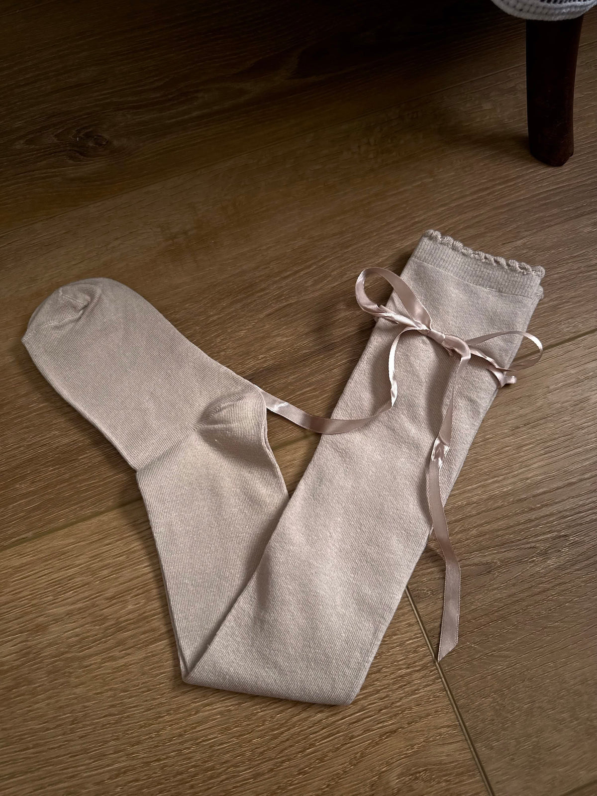 A pair of Historically Inspired Cotton Knee-High Socks with Satin Ribbon Lacing in oatmeal beige, pictured on a historic wooden cottage floor.