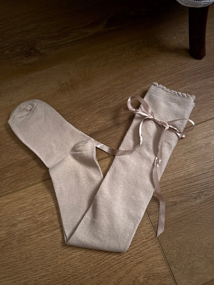 A pair of Historically Inspired Cotton Knee-High Socks with Satin Ribbon Lacing in oatmeal beige, pictured on a historic wooden cottage floor.