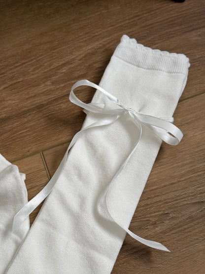 A pair of Historically Inspired Cotton Knee-High Socks with Satin Ribbon Lacing in Off-White, pictured on a historic wooden cottage floor.